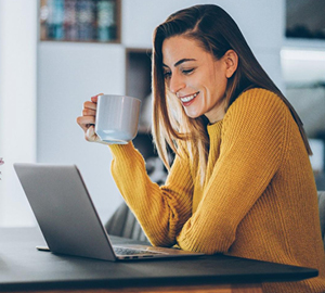 Young woman holding a cup of coffee smiling while working on her laptop.
