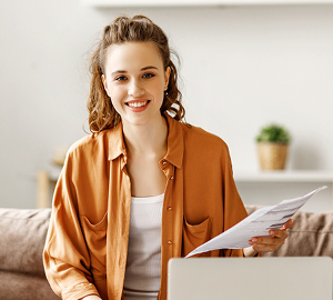 Young female smiling while holding paperwork looking at her laptop.