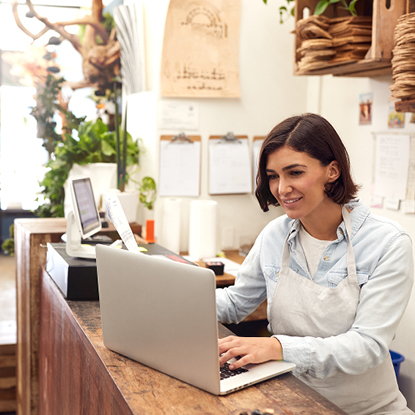 Shop worker wearing a white apron and smiling while using a laptop at her business.